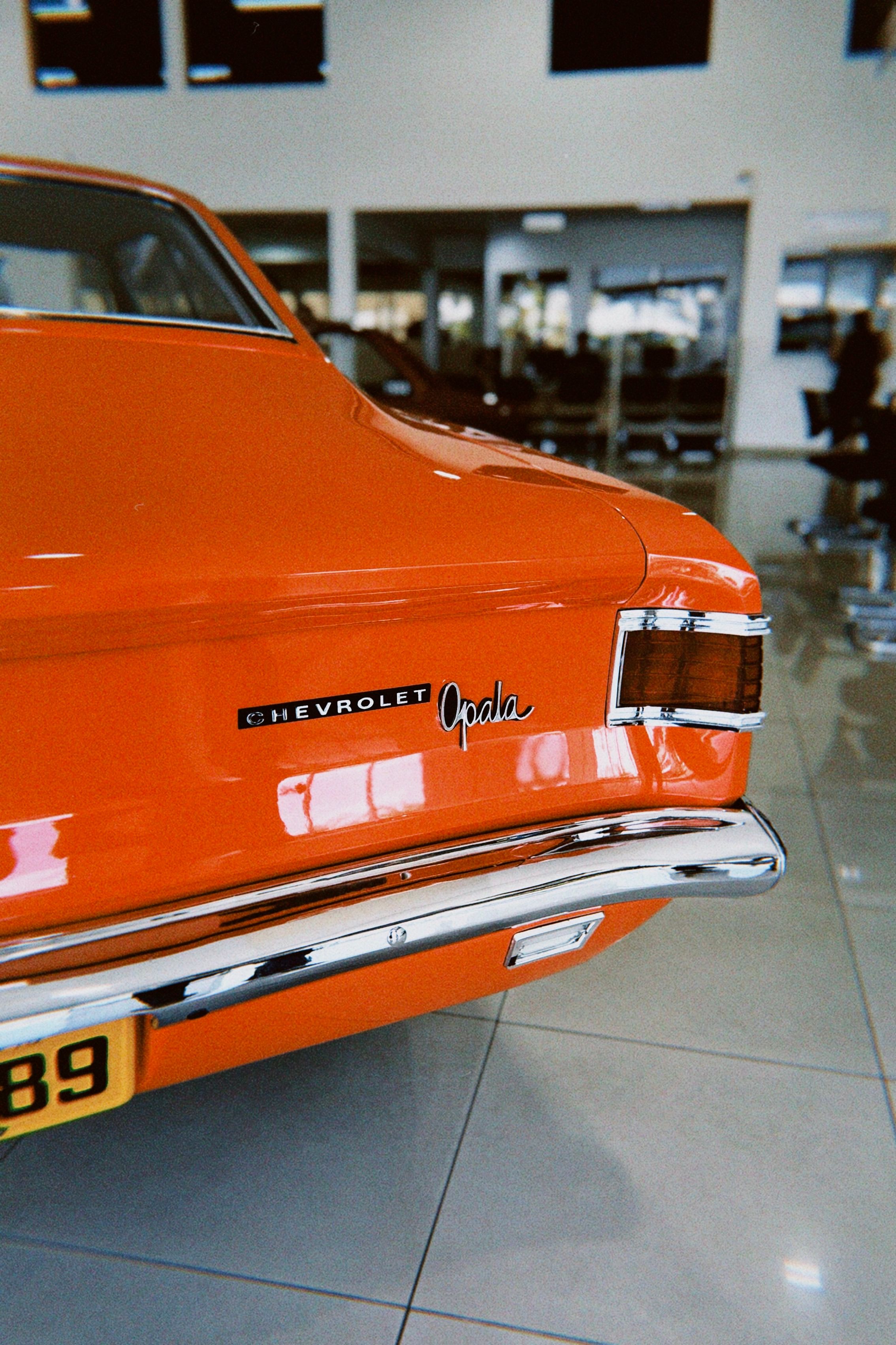 Close-up of a classic orange Chevrolet Opala's rear in a modern car showroom.