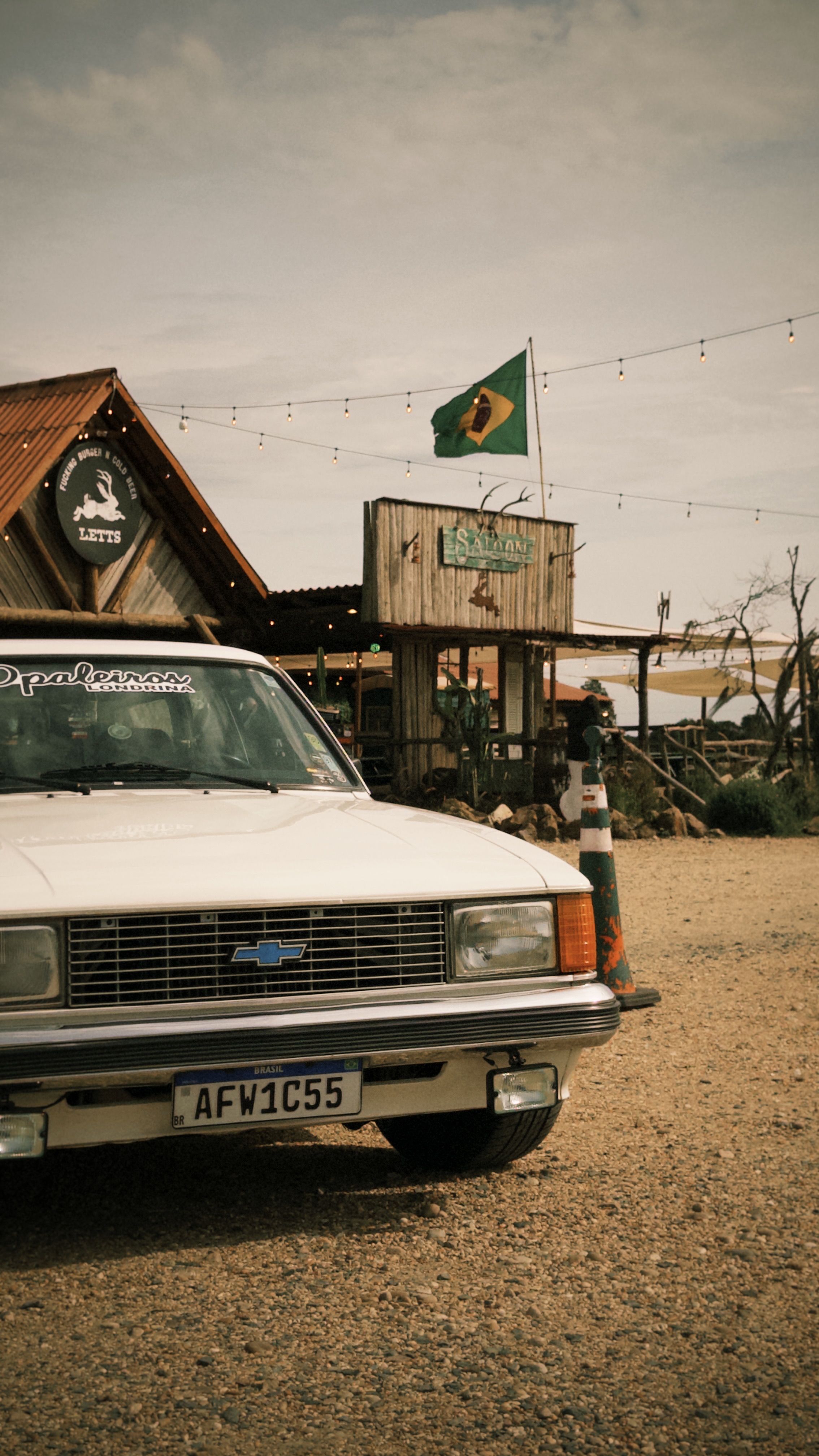 A vintage car parked at a rustic roadside stop in Brazil, featuring traditional architecture and flags.