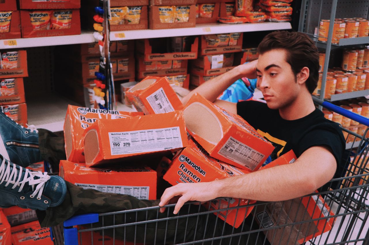 a man lounging in a shopping cart filled with maruchan ramen in a supe