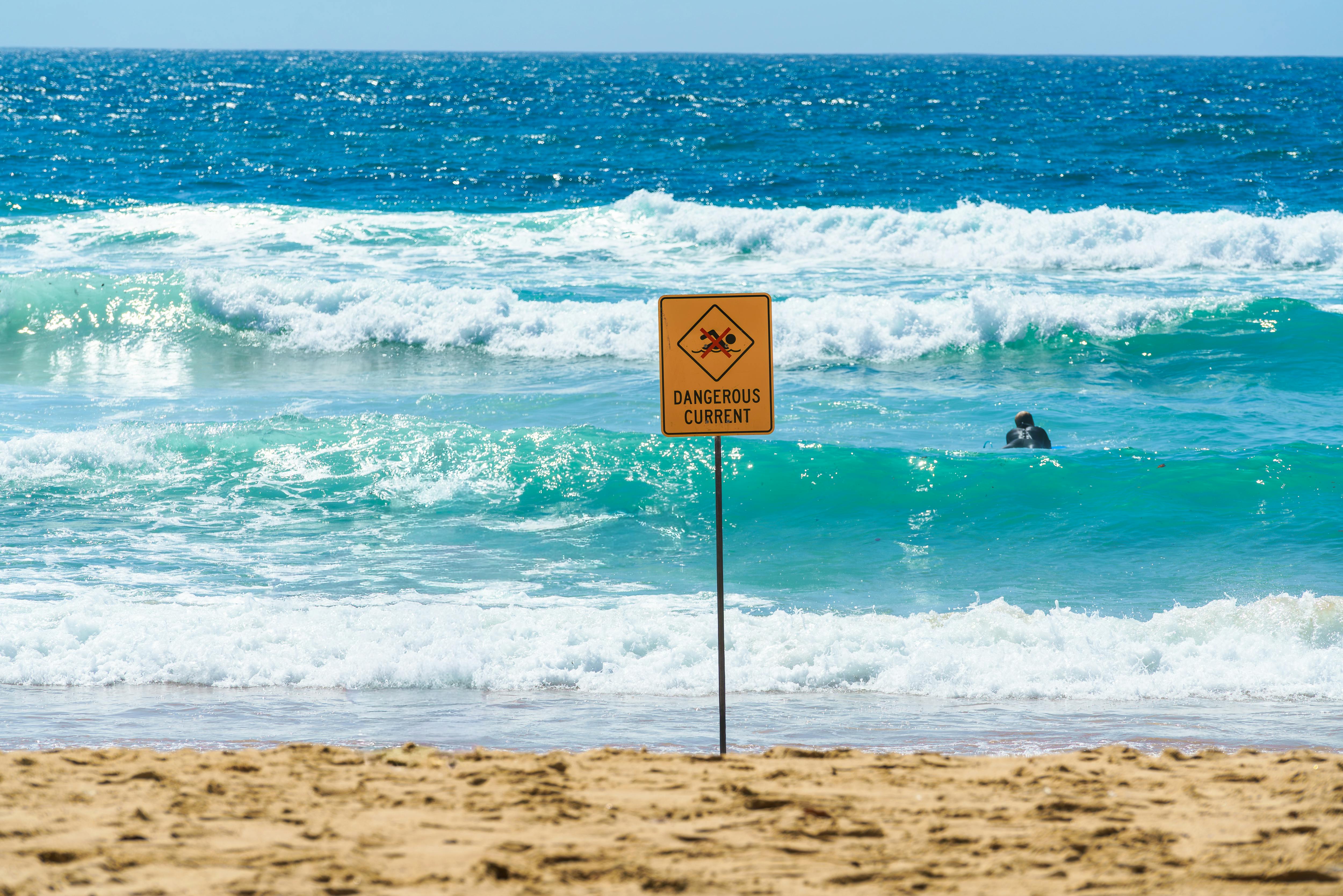 Beach scene with ocean waves and a warning sign for dangerous currents in Australia. | BocaVibe