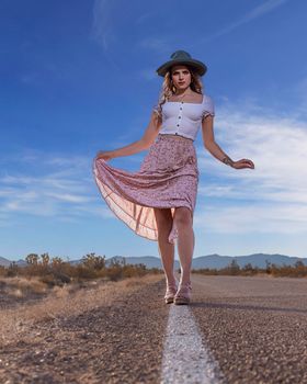 A woman in a hat and summer dress walking confidently along a desert road.