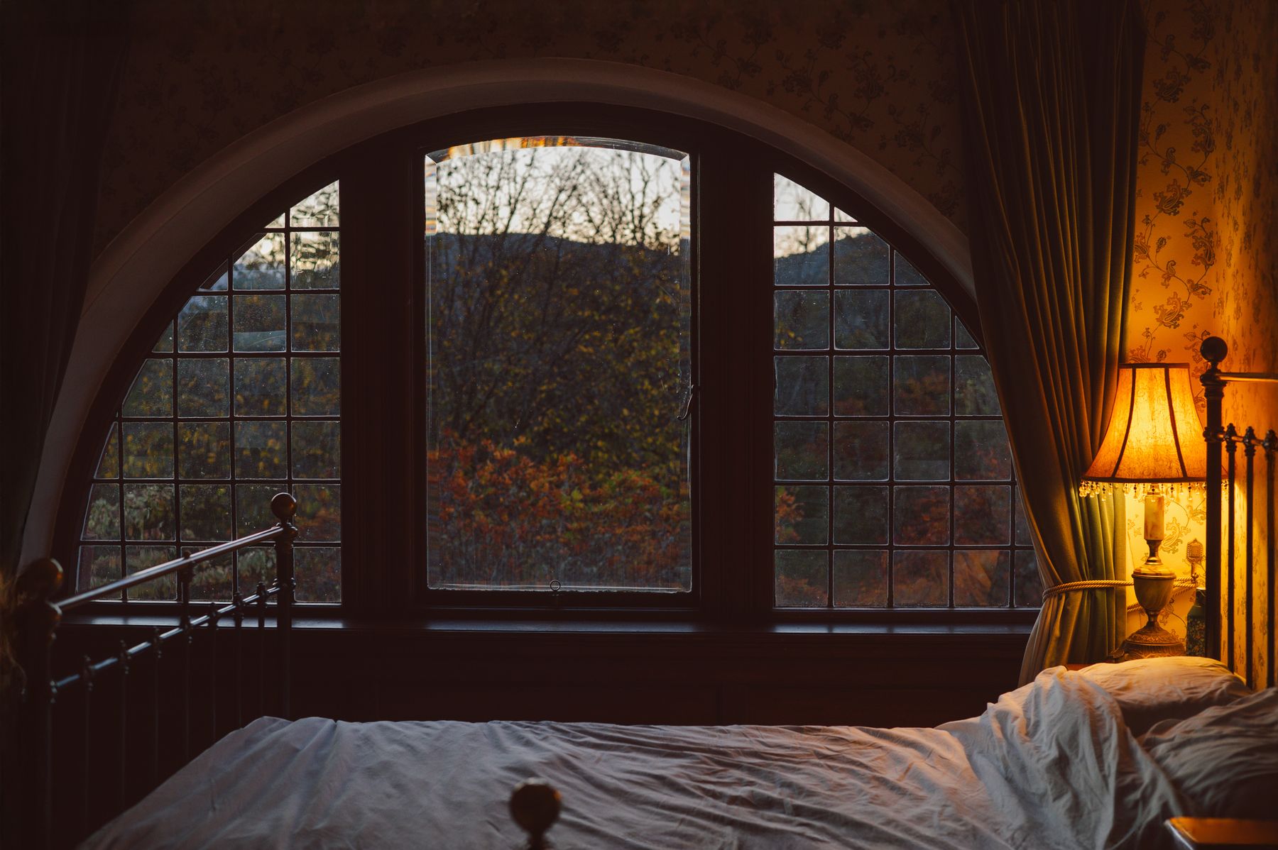 Metal bed beneath a broad arched window, patterned wall and warm fringed bedside lamp