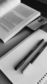 Black and white closeup of a study desk with an open book, grid notebook, and pens for a minimalist study scene.