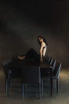 Elegant woman in stylish attire posing confidently on a minimalist black table in a dark, modern setting.