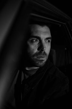 Dark, dramatic black and white portrait of a man sitting inside a car at night.