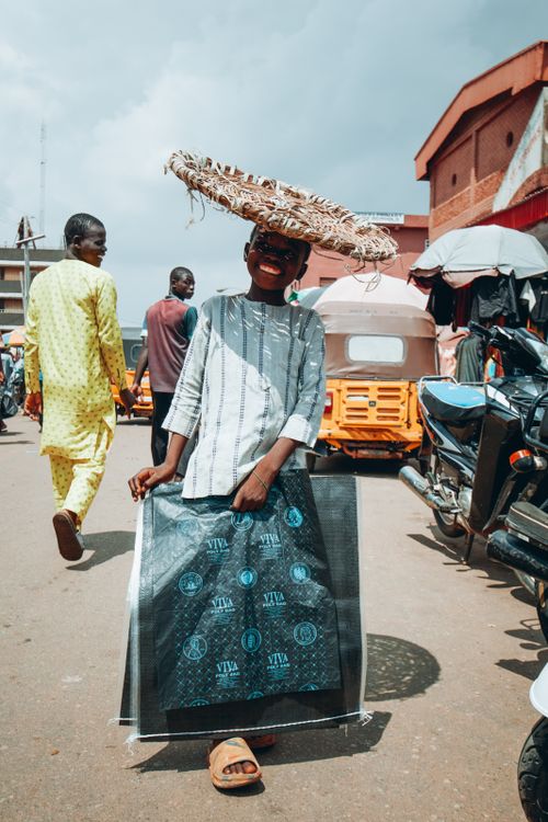 Free A young boy smiling under a large straw hat in a bustling market in Kaduna, Nigeria. Stock Photo