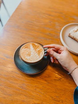 A hand gently holds a cappuccino cup on a wooden table, capturing a cozy coffee moment.