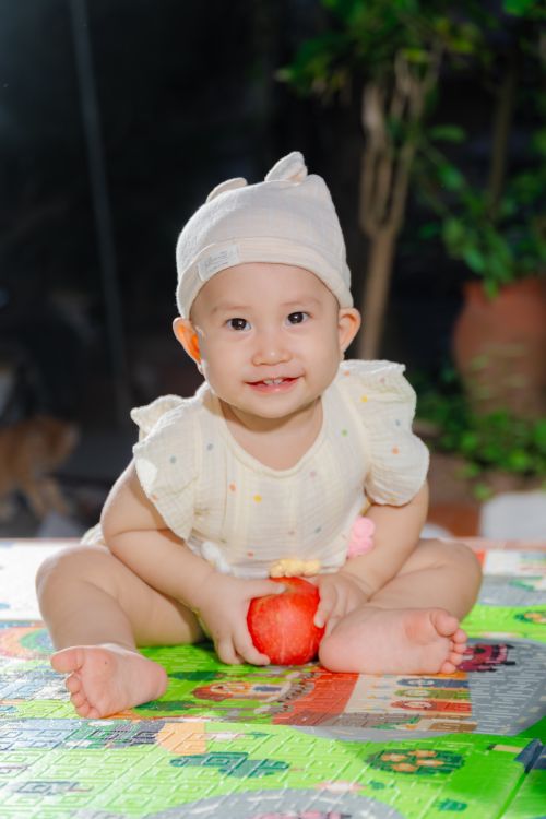Free Cute baby wearing a hat holding a red toy, sitting on colorful mat. Stock Photo