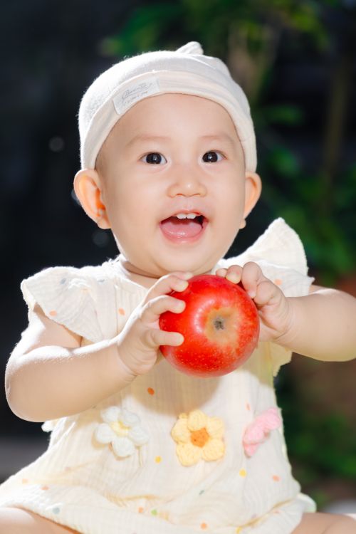 Free Joyful baby in a white dress holding a red apple, shot outdoors with soft lighting. Stock Photo