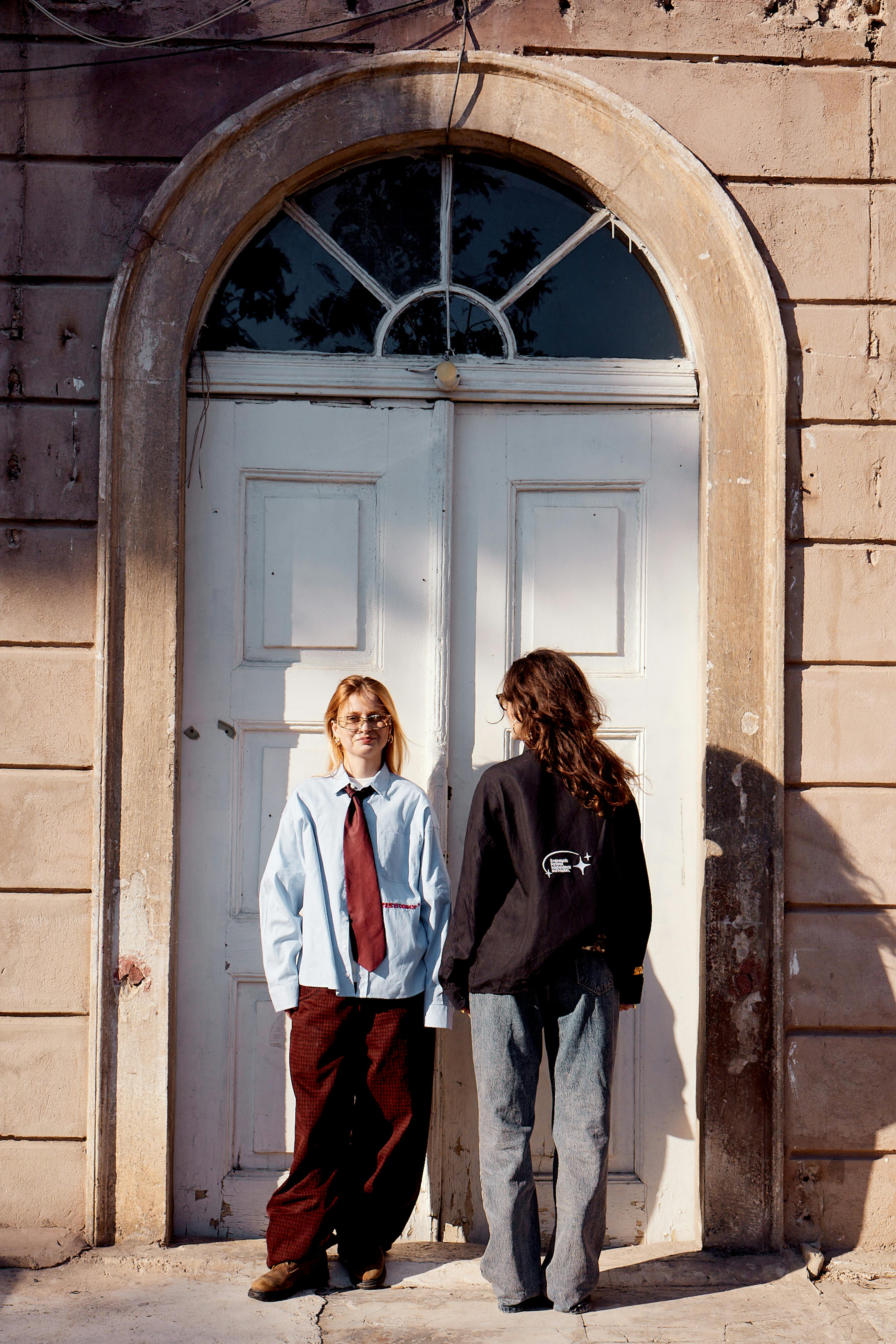 Two individuals in trendy attire stand in sunlight against a classic İstanbul doorway.
