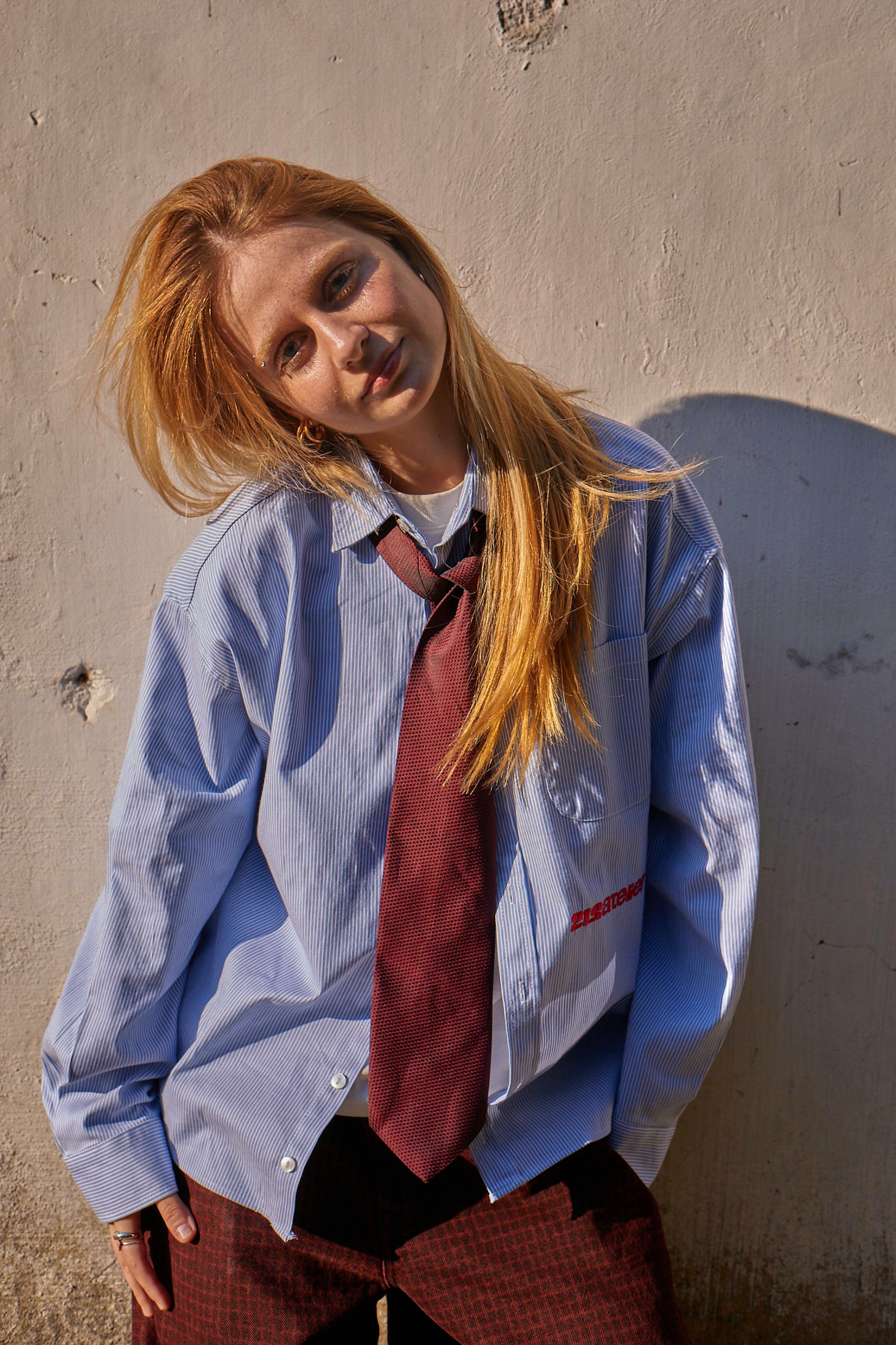 A stylish young woman poses in an oversized shirt and tie against a sunlit wall in Istanbul.