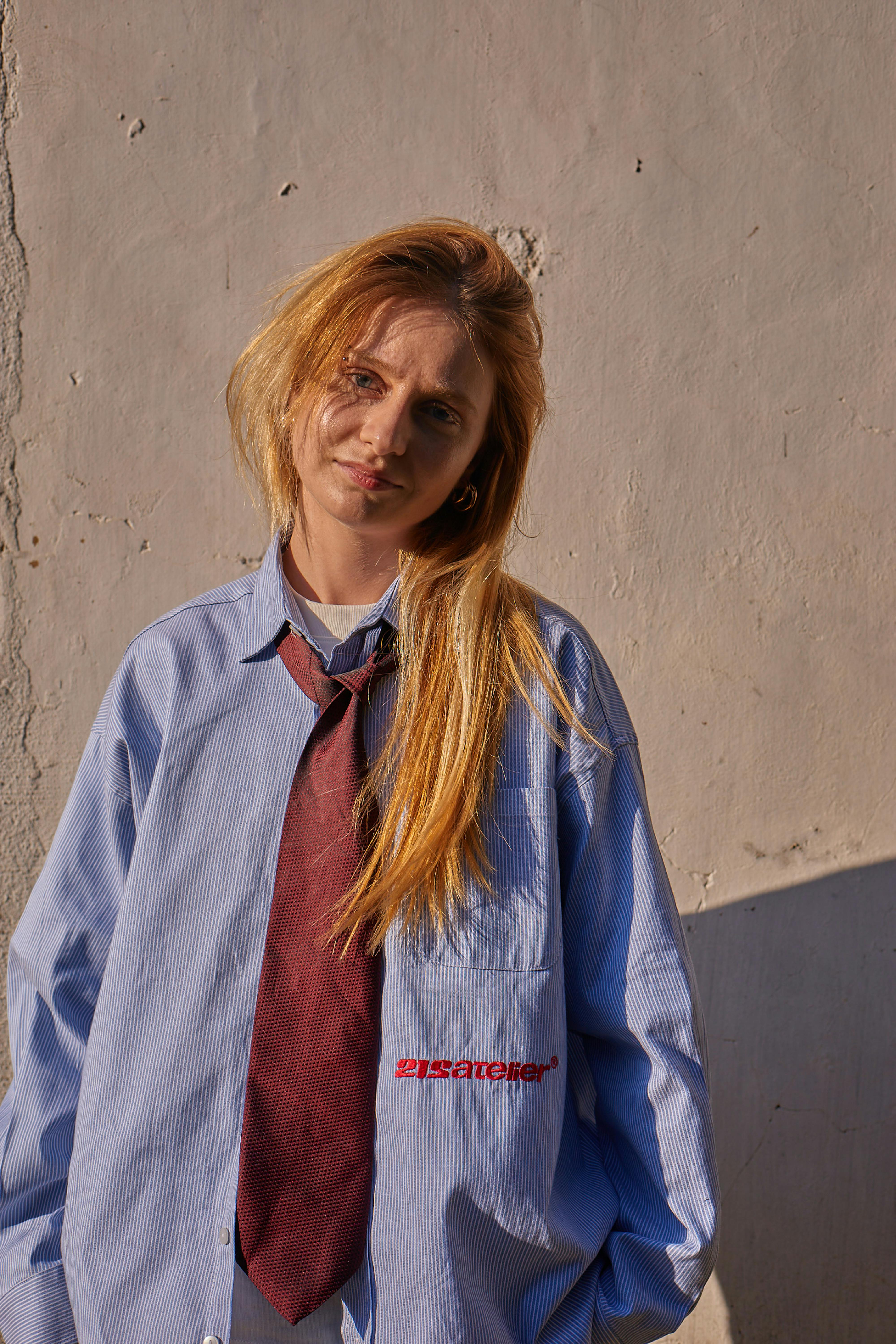 Portrait of a young woman in an oversized shirt and tie standing against a textured wall in Istanbul.
