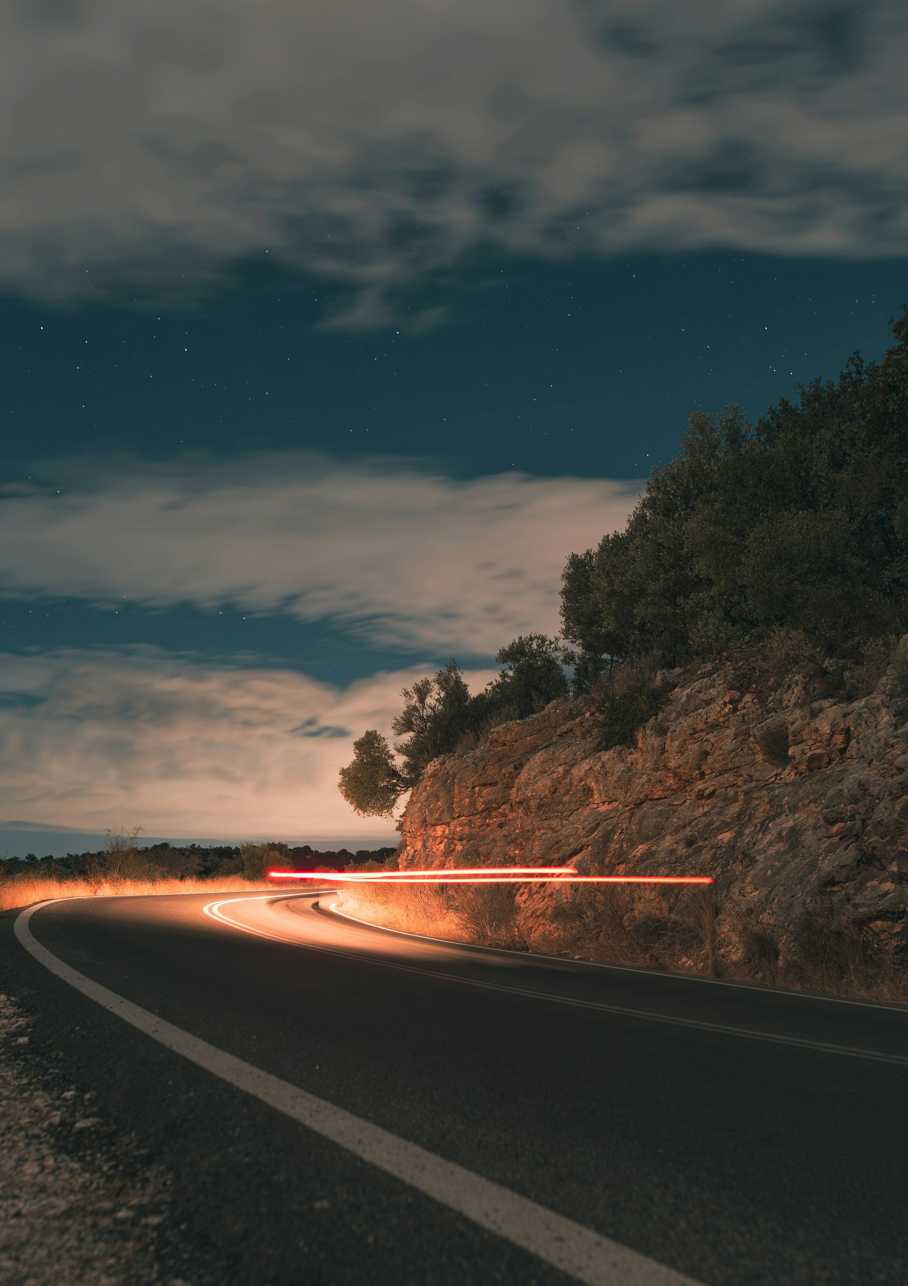 Curved mountain road at night with car light trails under a starry sky, showcasing night photography.