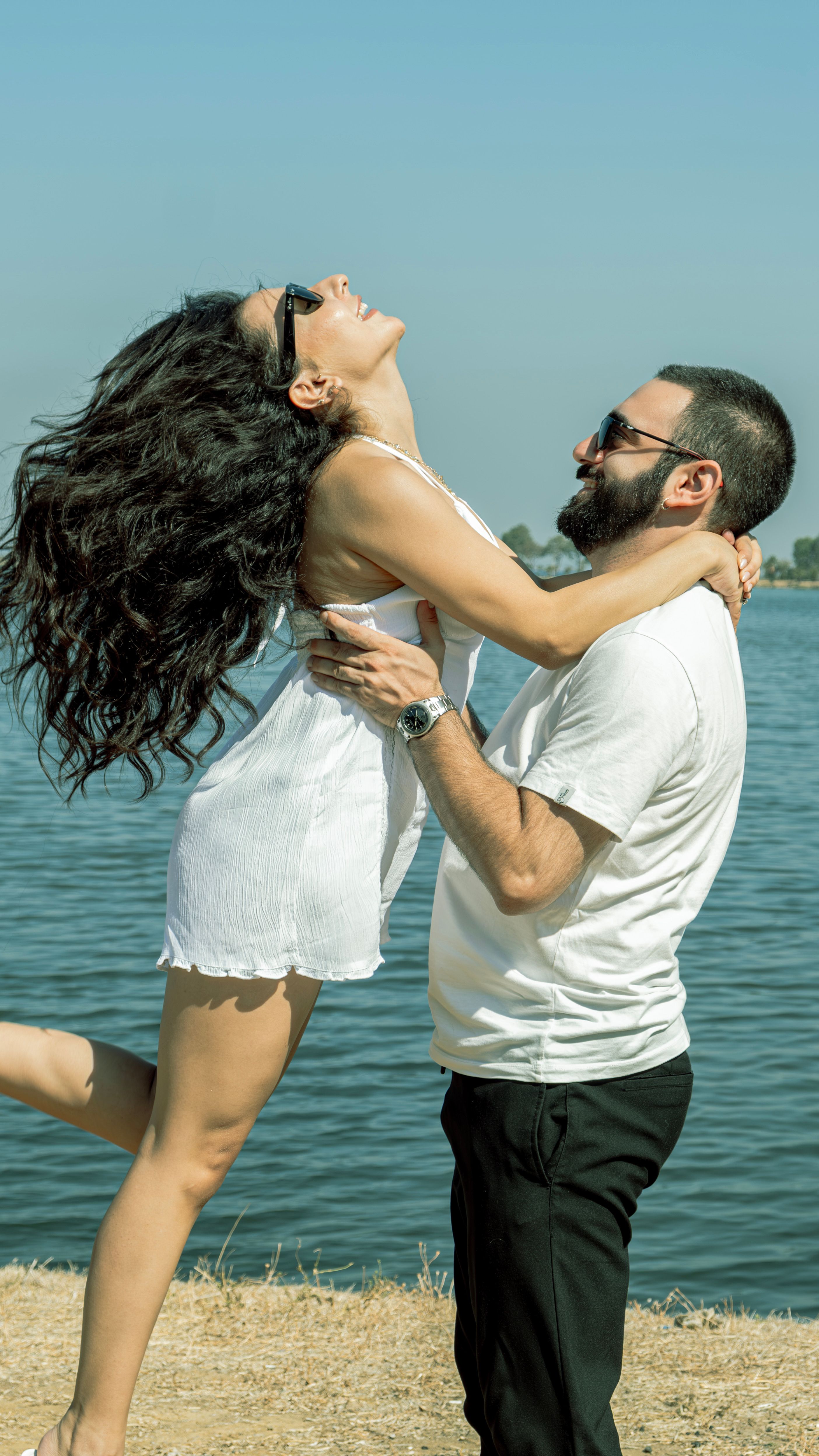 Happy couple laughing and embracing near a serene lake under clear blue skies.