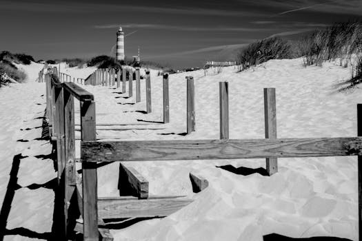 A striking black and white image featuring a sandy beach path leading to a distant lighthouse.