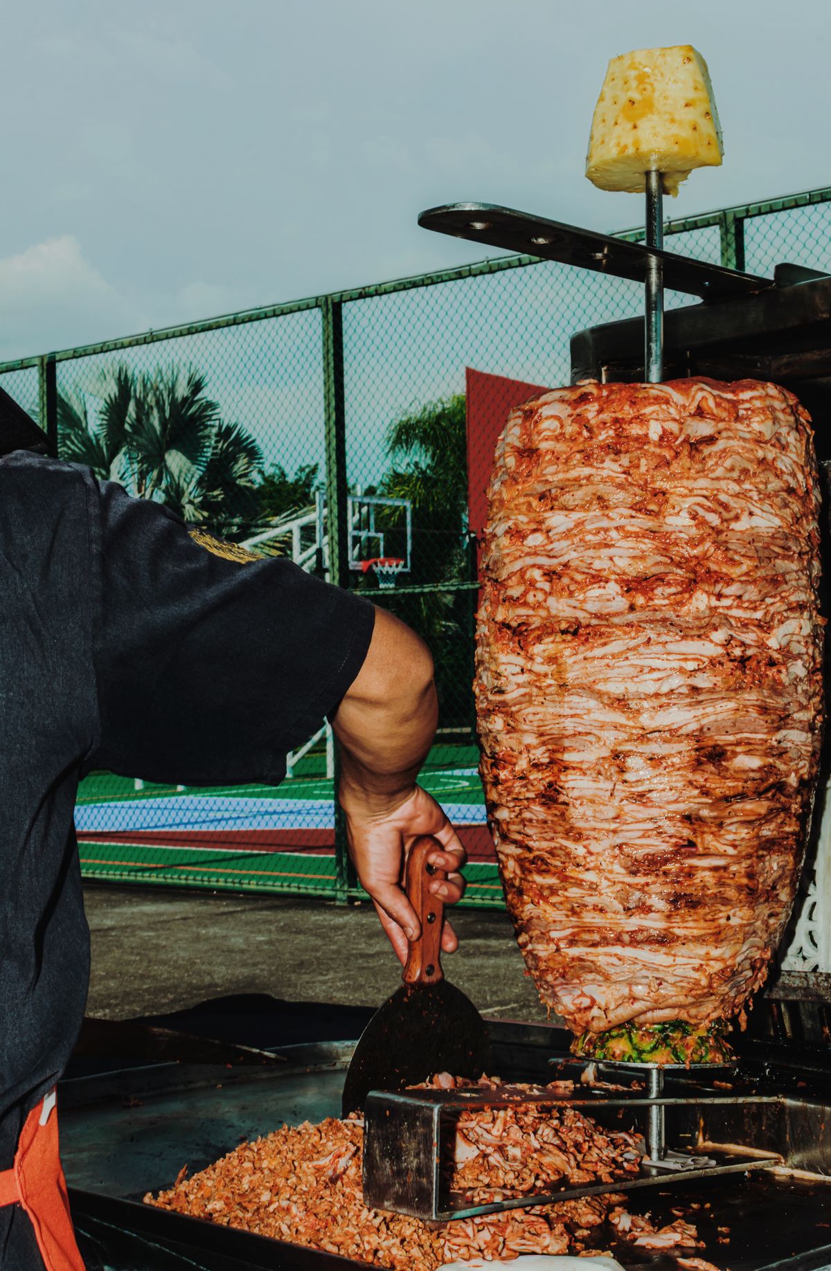 A vibrant scene of a street vendor slicing al pastor meat outdoors with a basketball court in the background.