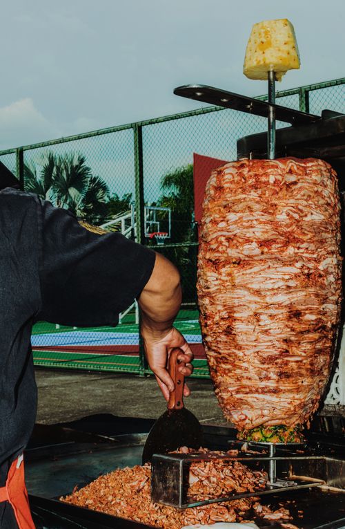 Free A vibrant scene of a street vendor slicing al pastor meat outdoors with a basketball court in the background. Stock Photo