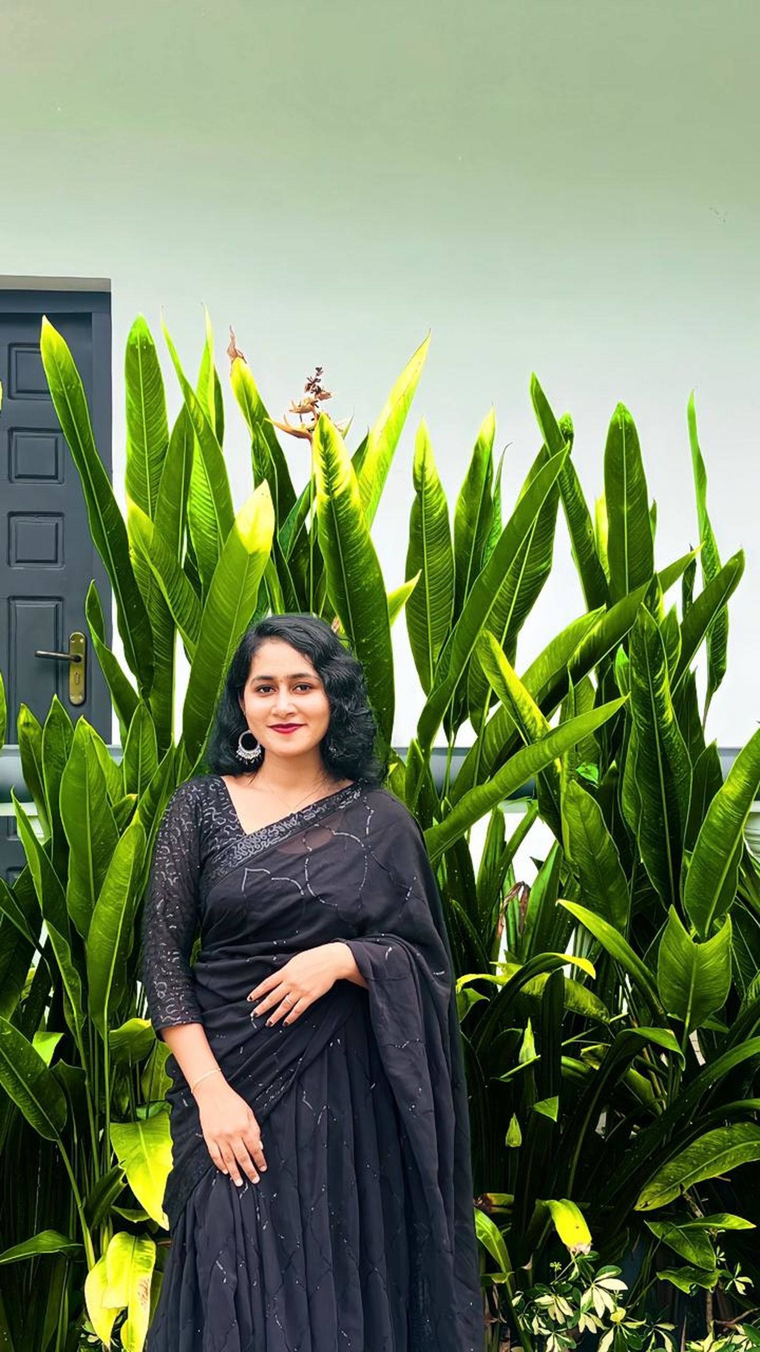 Woman in black saree posing against lush green plants by a modern door.