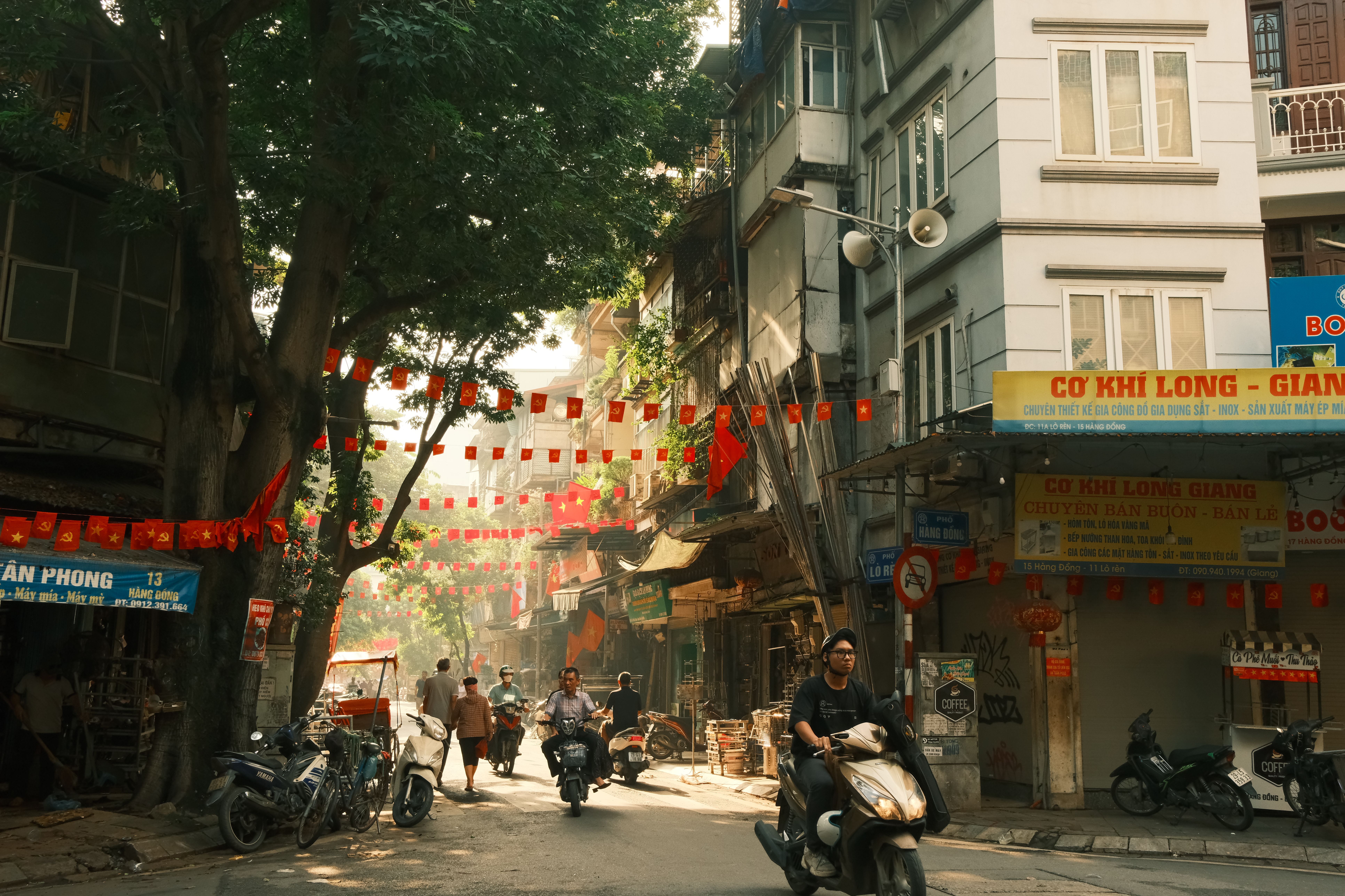 Asian city street bustling with motorbikes, vibrant red flags, and local shops in warm daylight.