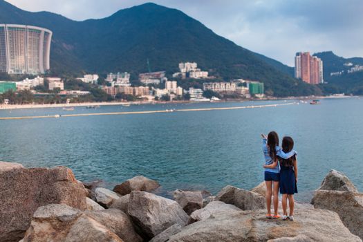 Two girls embrace and point across the water towards buildings on Hong Kong Island.