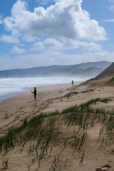 Two people fish along a serene Australian beach with scenic dunes and sweeping clouds.
