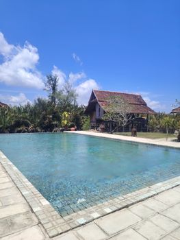 A tranquil poolside scene with a traditional house under a bright blue sky.