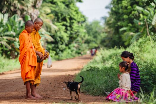 Monks in vibrant robes receive alms from a family on a rural path in Battambang, Cambodia.