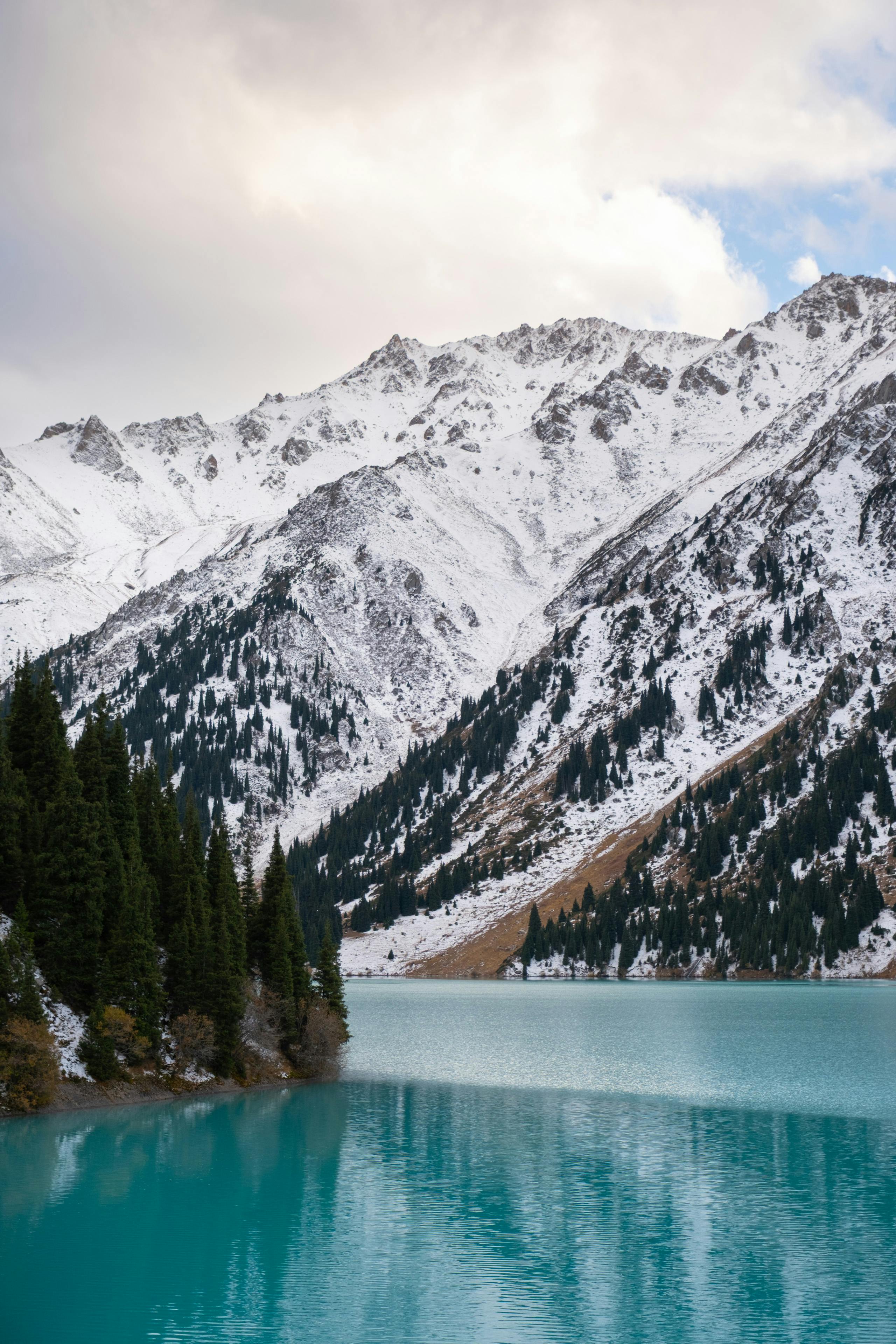 Snow-covered mountains reflecting in Big Almaty Lake, Kazakhstan, under a partly cloudy sky.