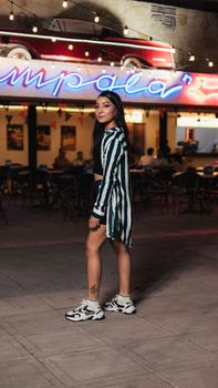 Fashionable young woman in striped attire poses in front of a vibrant cafe with neon lights at night.