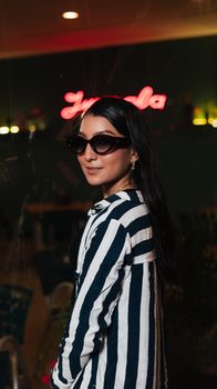 Stylish woman in sunglasses and striped shirt at a cozy cafe with neon lights.
