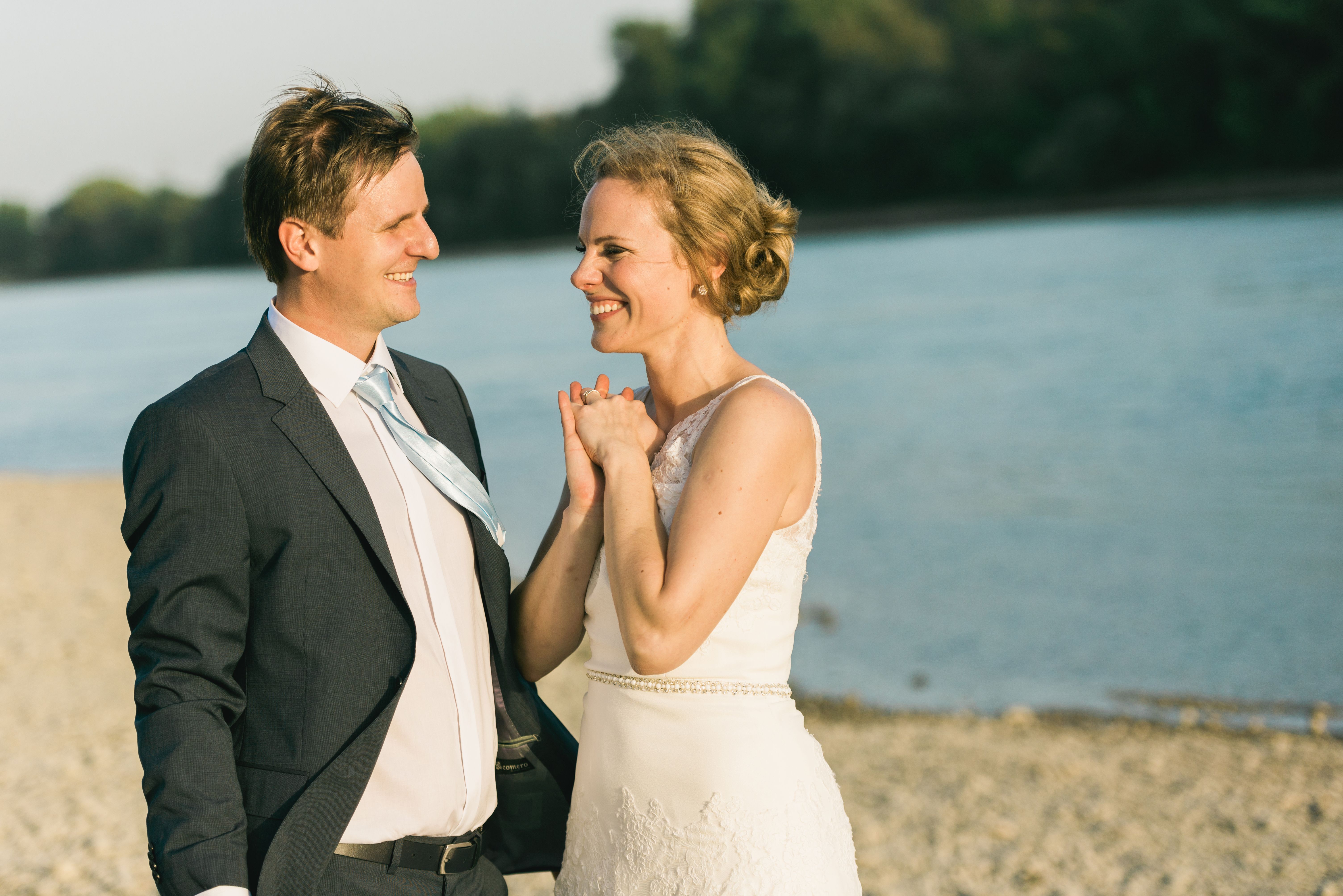 Bride and groom in elegant attire sharing a joyful moment by the riverside on a sunny day.