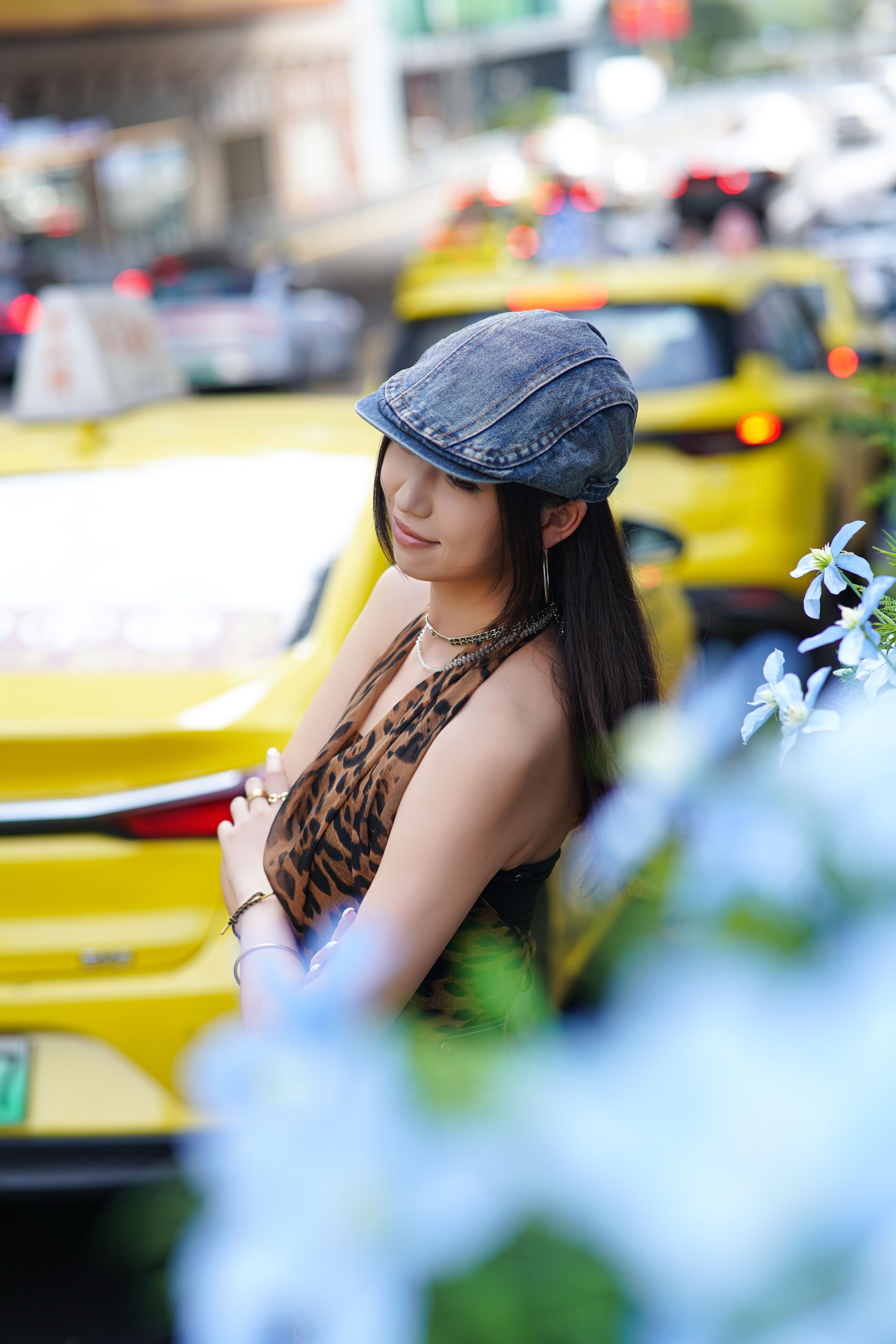 Stylish woman posing outdoors next to city traffic with taxis, wearing a cap and leopard print top.
