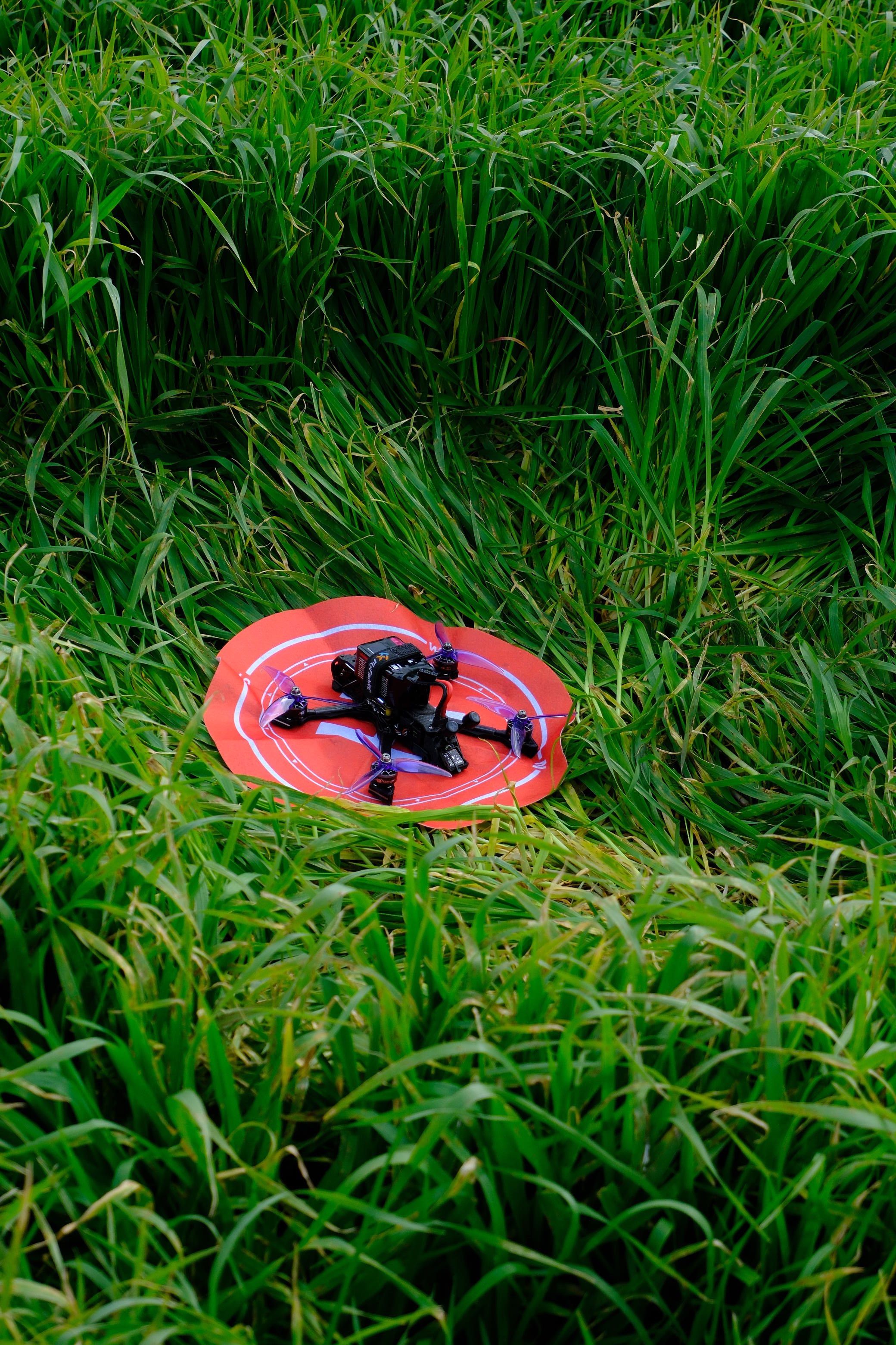 A drone rests on a bright red landing pad amidst lush green grass, outdoors.