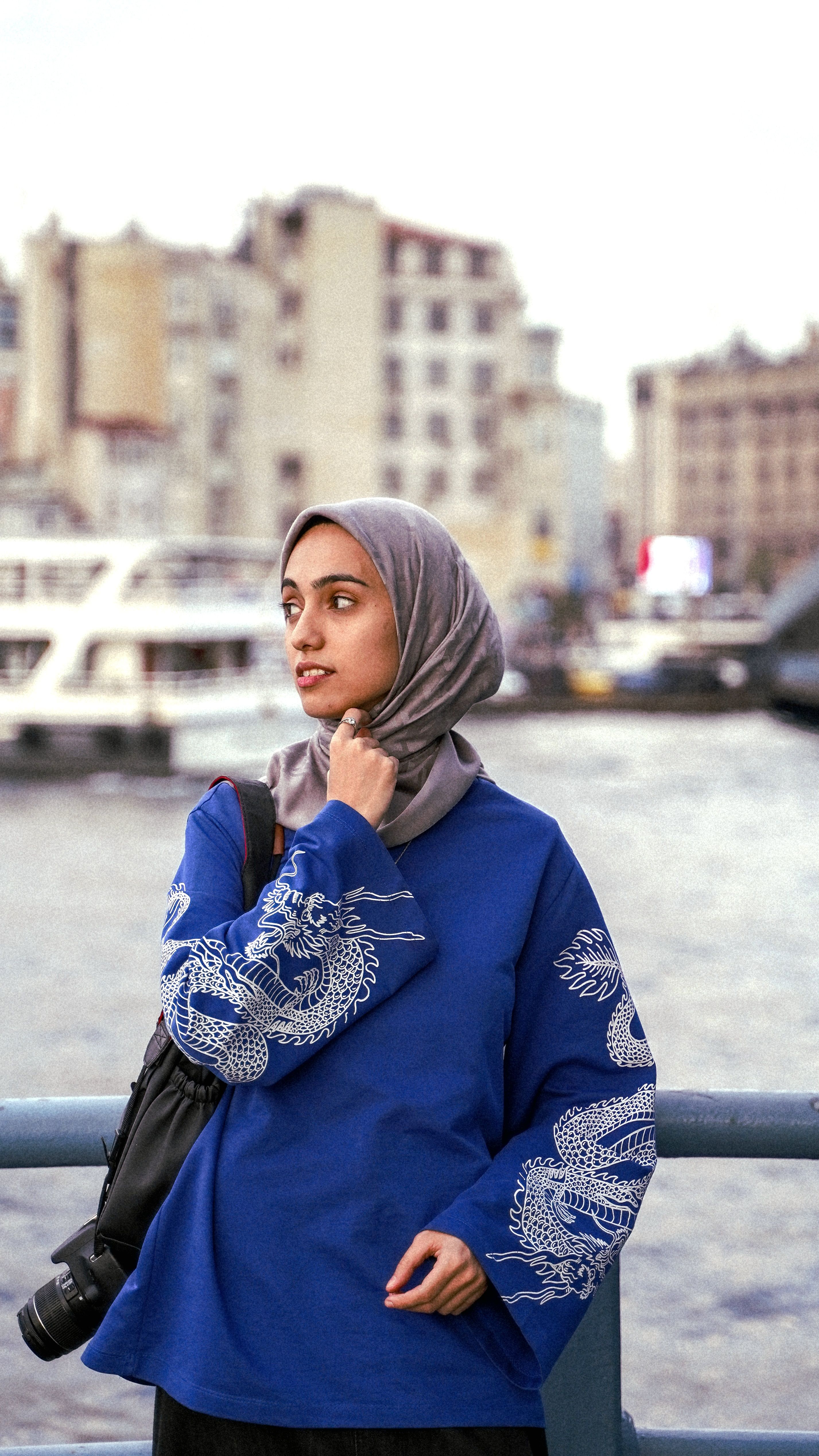 Portrait of a woman in a blue hijab and sweater near a city waterfront, expressing modern fashion.