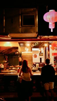 Colorful night market scene with food vendor and customers under glowing lantern.