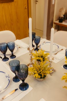 A beautifully arranged dining table with blue glassware, white candles, silver holders, and yellow flowers.