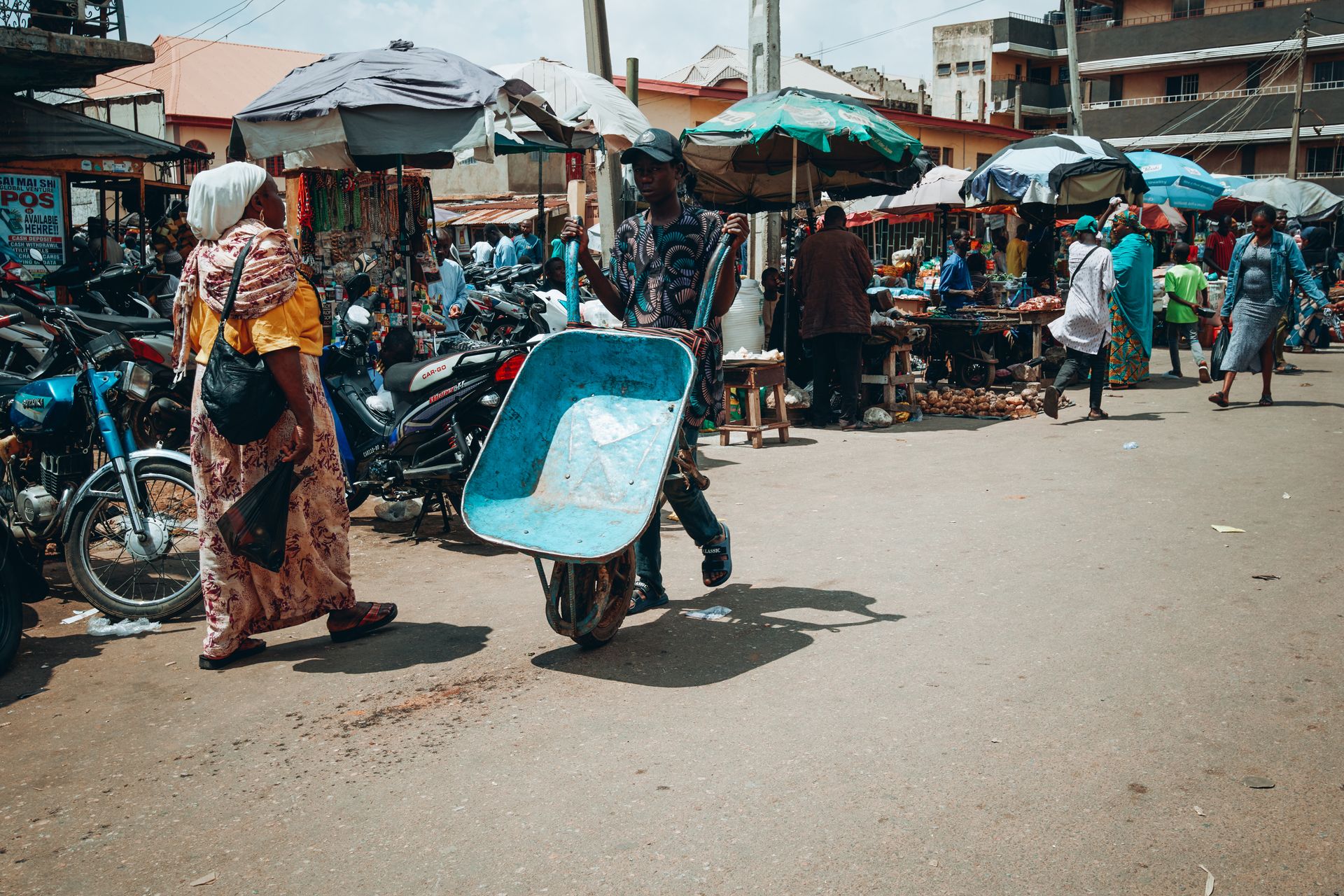 Busy kaduna central market vibes in Nigeria! 🚀 A guy's got his wheelbarrow game strong, transporting goods through the bustling streets while folks shop under colourful umbrellas. Motorbik...