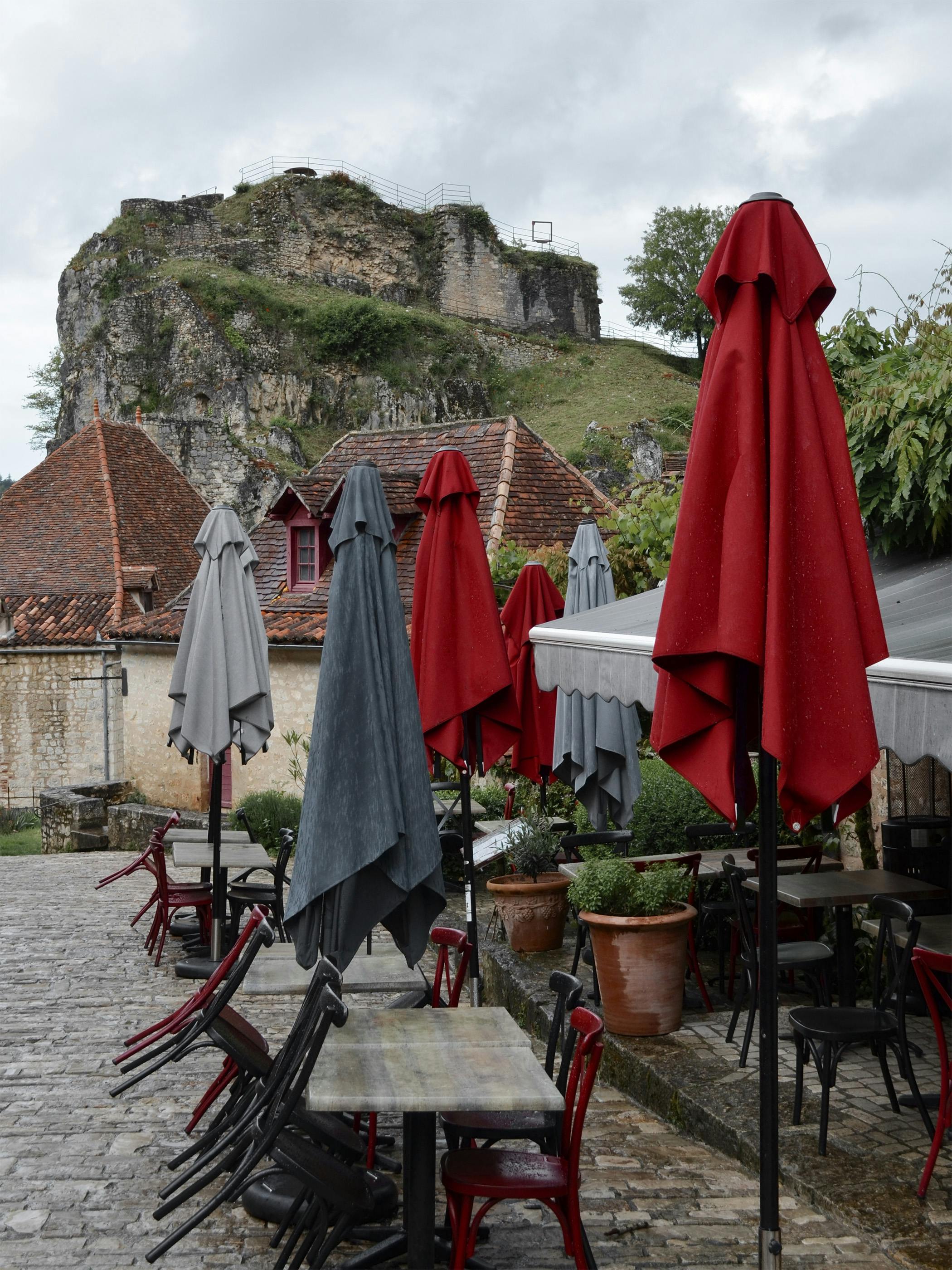 Quaint outdoor cafe in Saint-Cirq-Lapopie, France, with closed umbrellas and stone architecture.