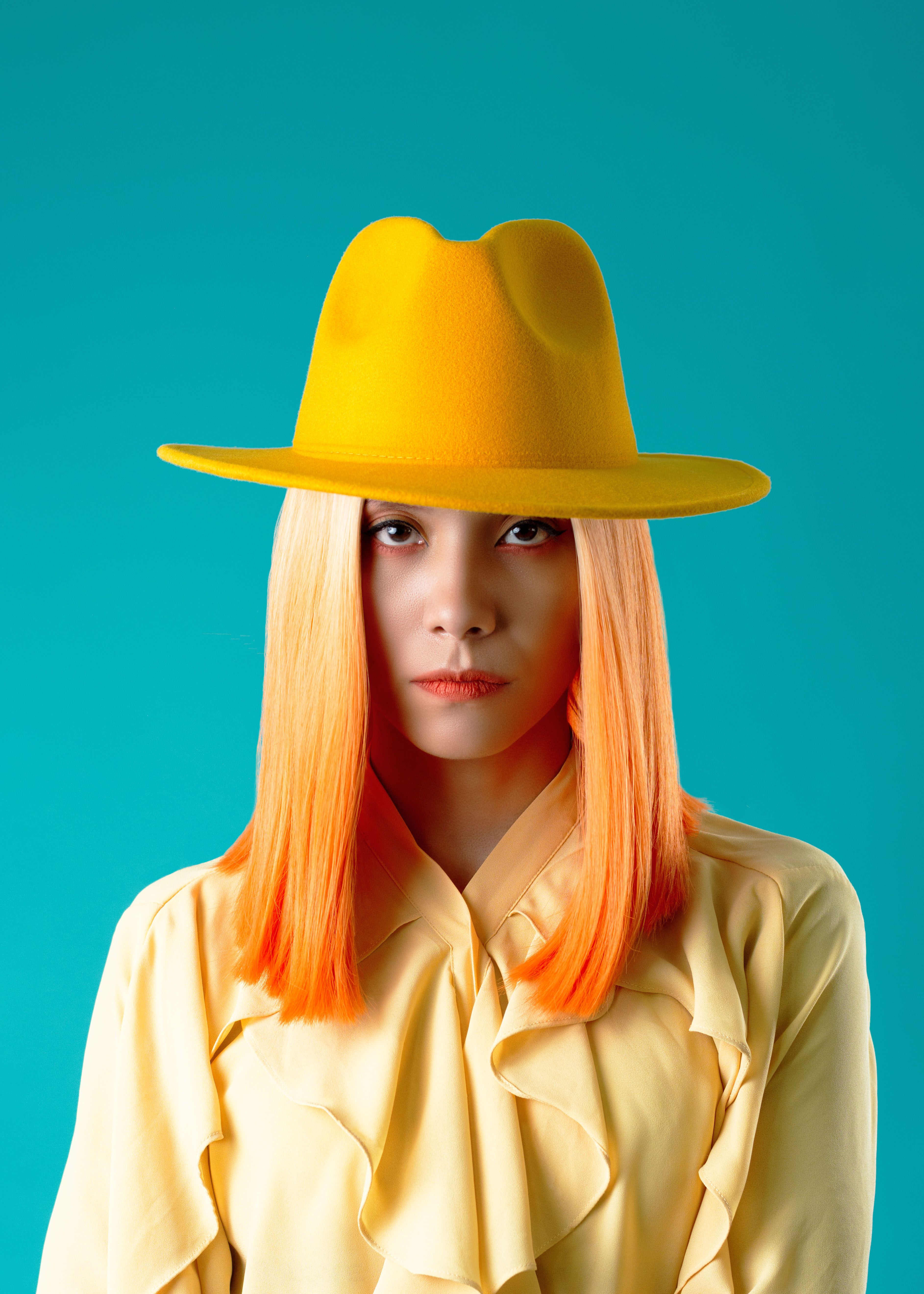 Vibrant portrait of a woman with orange hair and yellow hat against turquoise background.