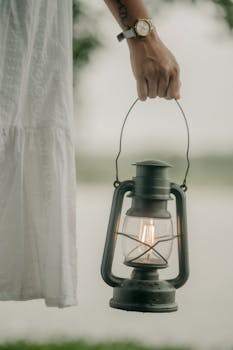 A person in a white dress holds a vintage lantern with a warm glow outdoors.