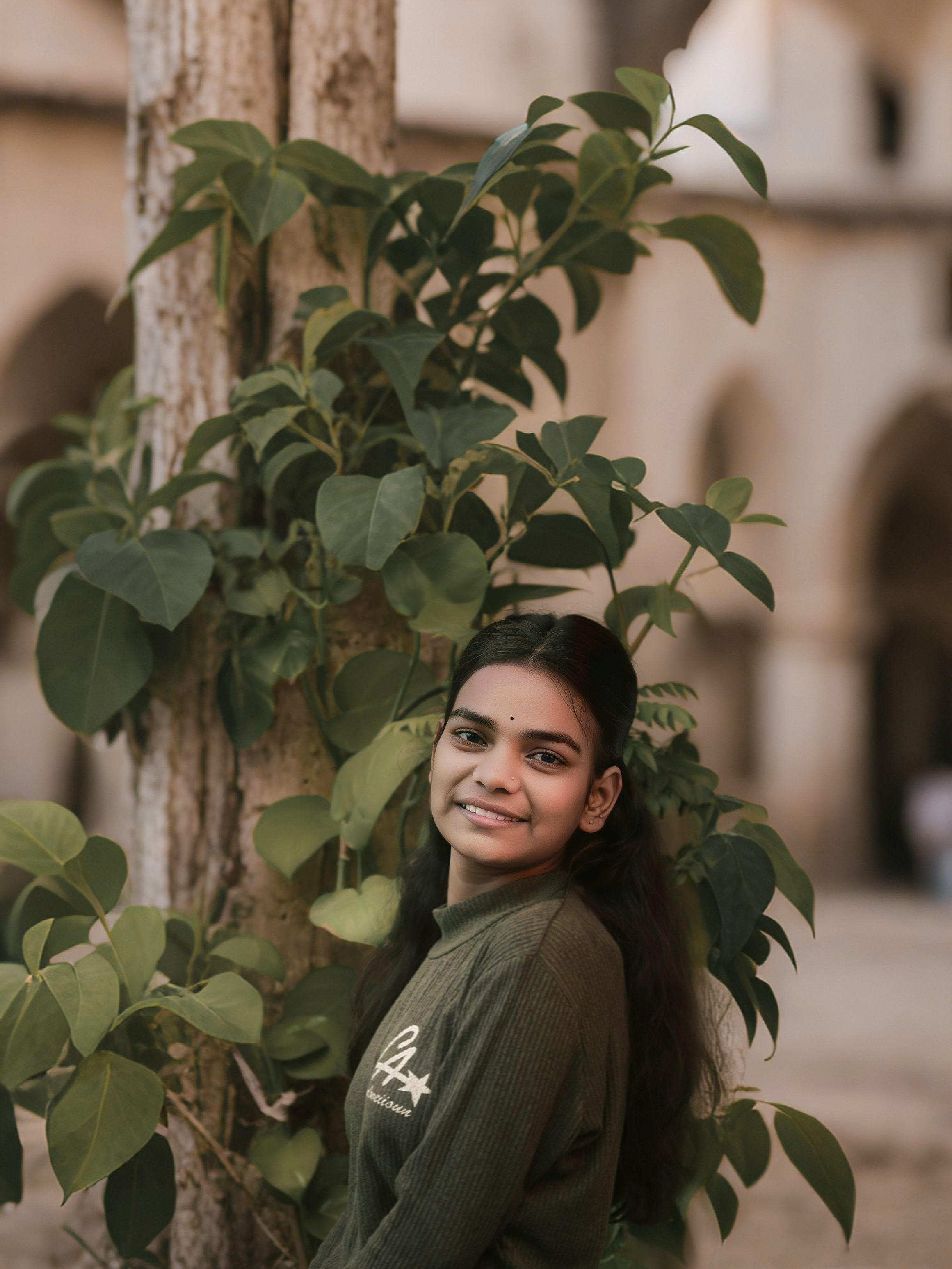 A young woman smiling and posing beside a tree with lush green leaves in a rustic architectural setting.