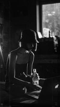 Black and white photo of a woman using a laptop by a window, creating a moody silhouette.
