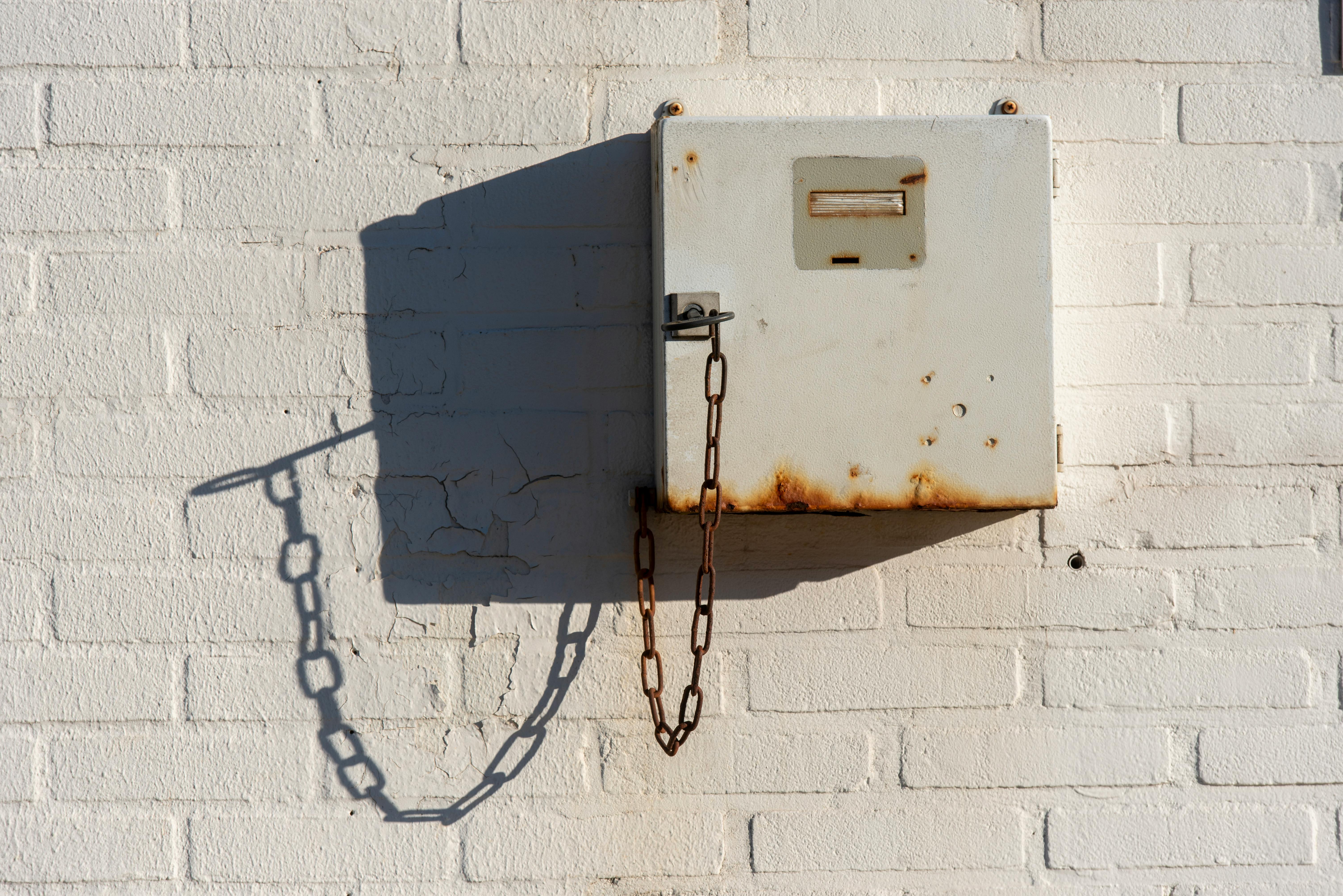 Free Rusty metallic utility box with chain on a white brick wall capturing shadows and texture. Stock Photo