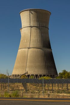 A cooling tower at a power plant surrounded by a fence and trees; under a bright blue sky.