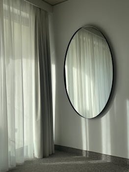 A serene minimalist room featuring a round wall mirror and sheer curtains.