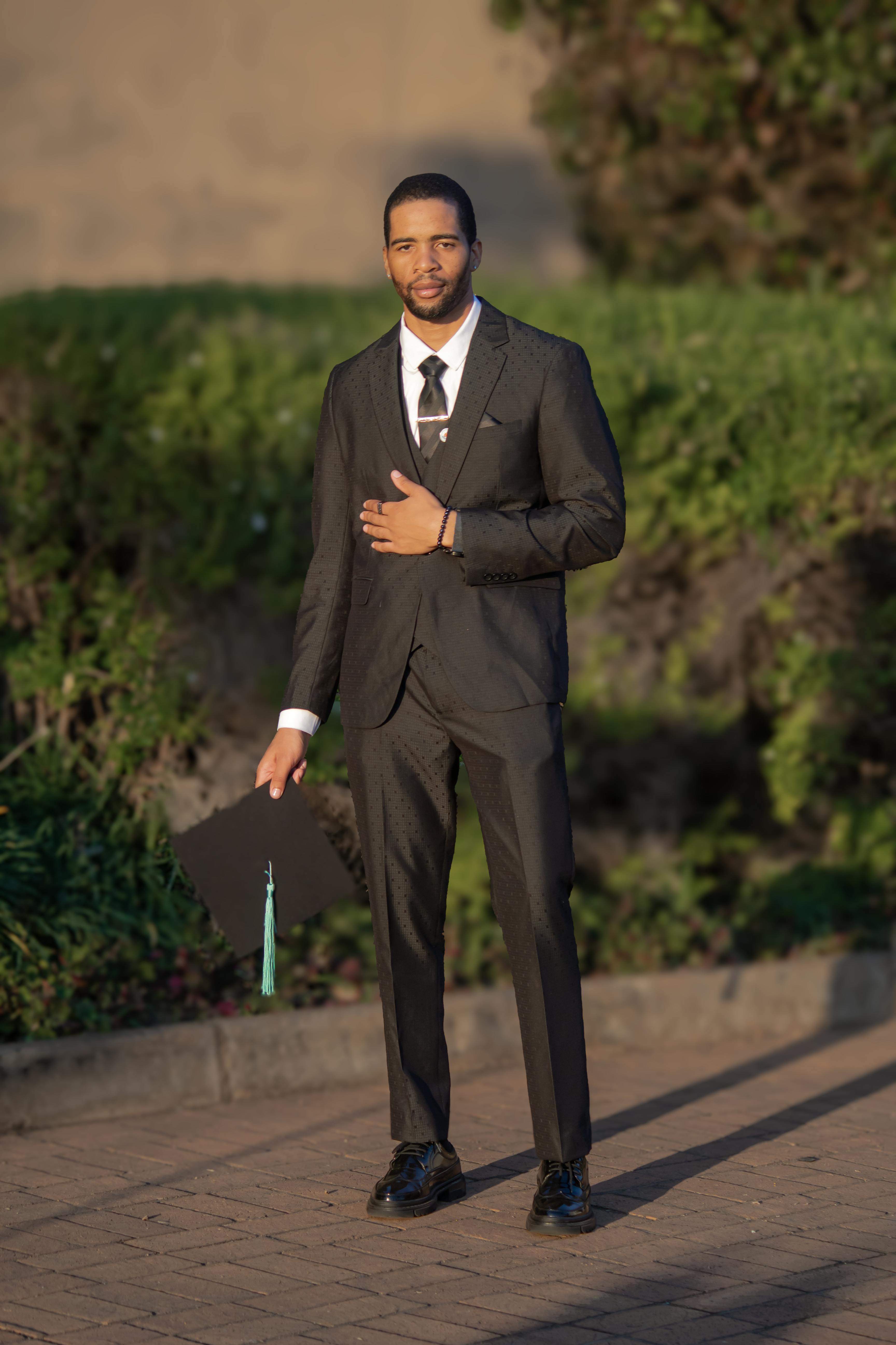 grátis Um jovem de terno formal segura um chapéu de formatura ao ar livre, comemorando conquistas acadêmicas. Foto profissional