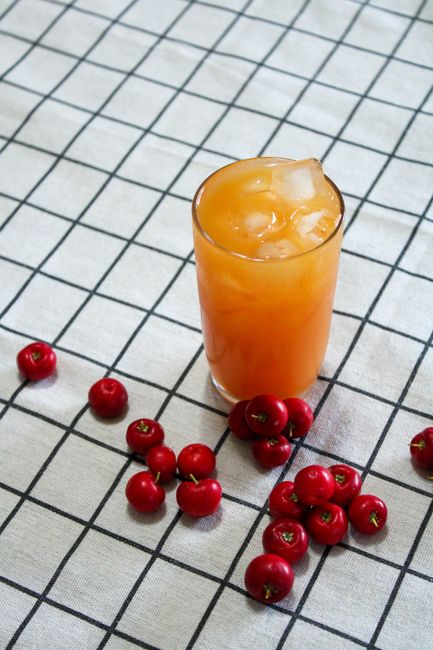 Refreshing acerola juice with ice surrounded by ripe acerola berries on a checkered tablecloth.