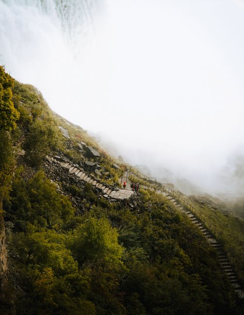 Misty waterfall view with people hiking on a lush, green pathway.