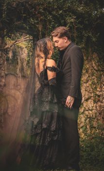 A couple dressed in gothic attire embracing in a natural setting, expressing deep affection.