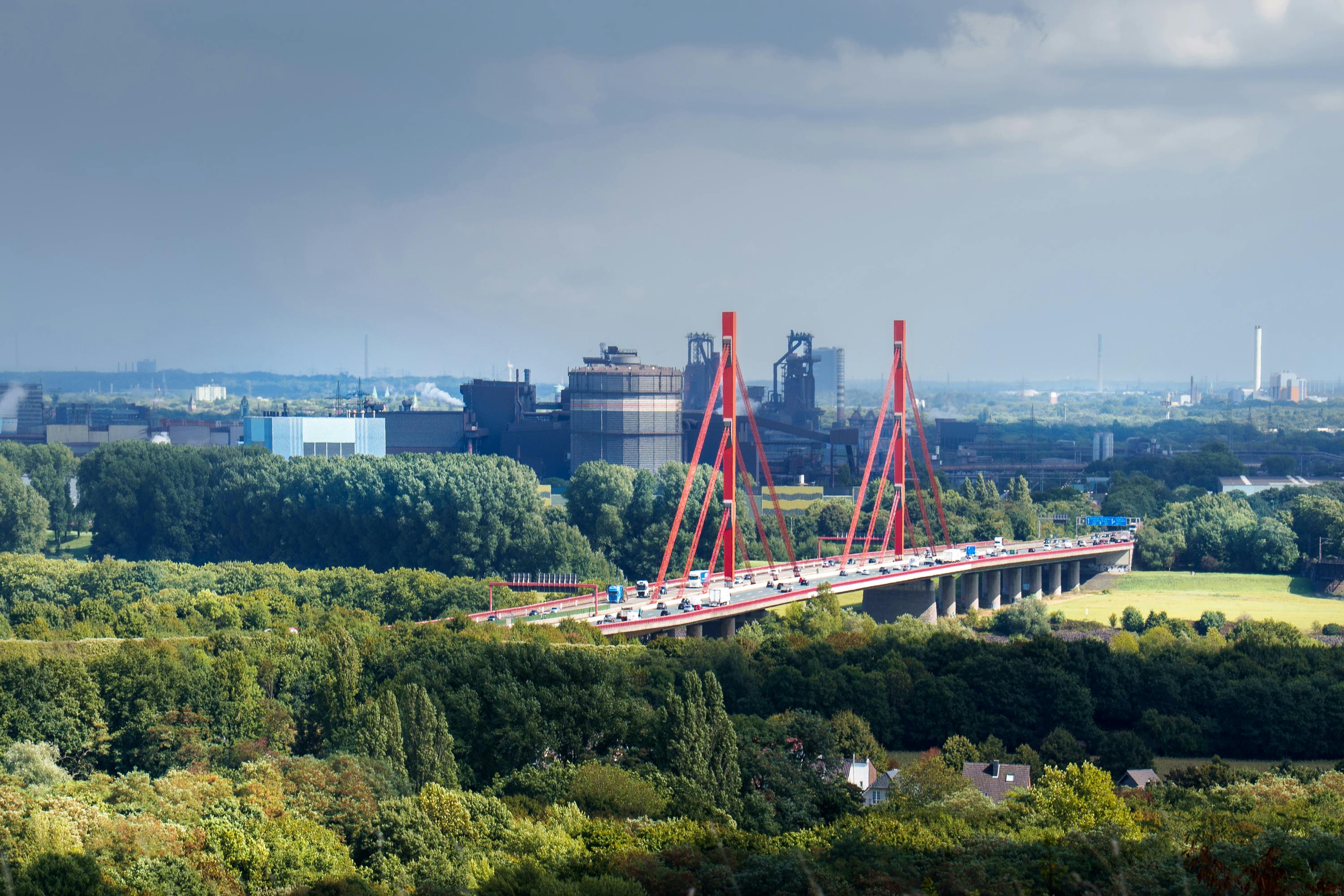 Aerial shot of an industrial area with a red bridge amidst greenery and urban skyline.